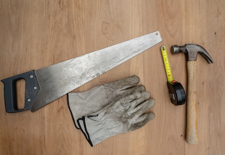 Hand saw, work gloves, tape measure, and hammer laid out on a wood surface, the kind of tools and hardware stocked at Bomgaars